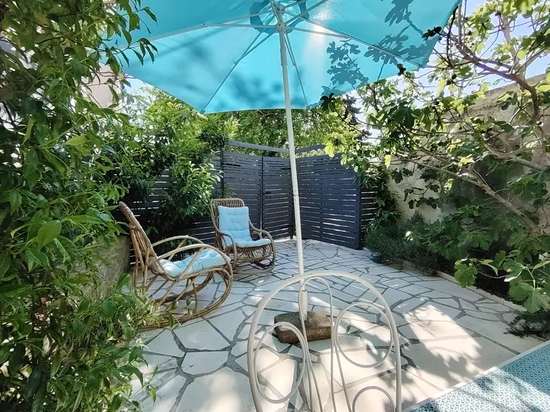 Gepflasterte Terrasse von Casa Bella mit türkisem Sonnenschirm, zwei Rattan-Schaukelstühlen und Feigenbaum