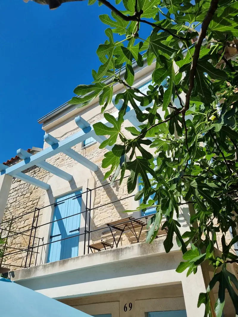 Casa Bella: Natursteinfassade mit weißer Pergola, blauen Fensterläden und Feigenbaum vor blauem Himmel